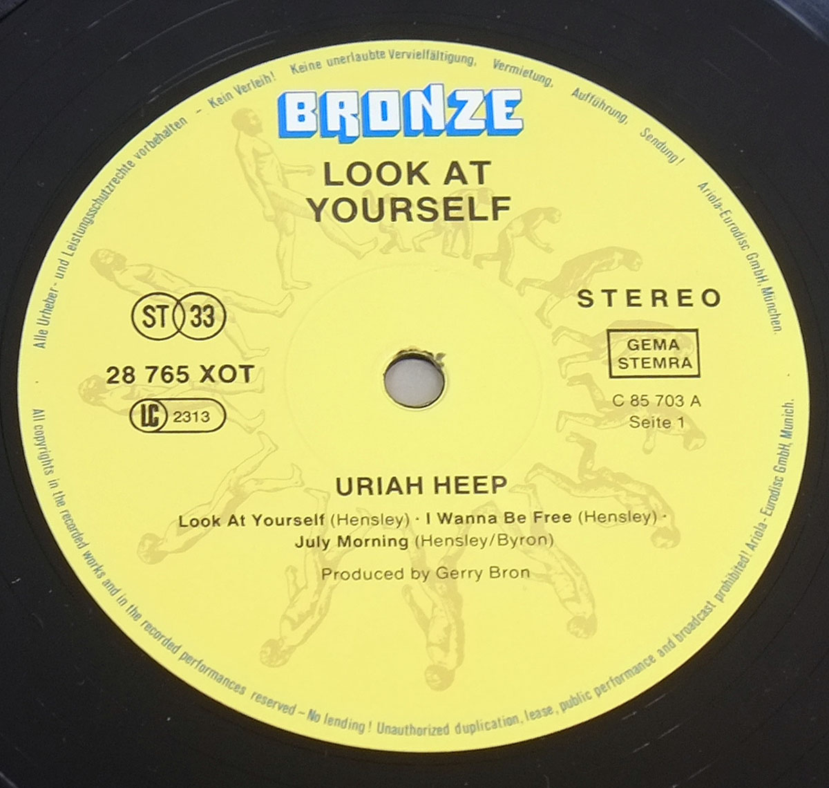Close-up of a yellow vinyl record label for Uriah Heep’s Look at Yourself, released on Bronze Records. The label features the blue ‘Bronze’ logo at the top, with black text listing the album title, band name, catalog number (28 765 XOT), and tracklist for Side 1: ‘Look At Yourself,’ ‘I Wanna Be Free,’ and ‘July Morning.’ The label also includes rights societies GEMA and STEMRA, a ‘Seite 1’ marking, and a circular pattern of running human figures in the background. Produced by Gerry Bron.